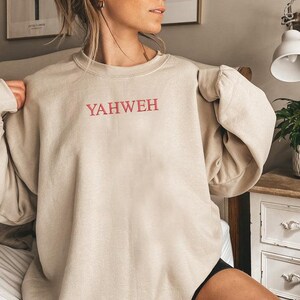 May include: A beige crewneck sweatshirt featuring the word YAHWEH embroidered in red capital letters across the center of the chest. The garment has long sleeves with ribbed cuffs and a ribbed neckline, constructed from a solid-colored fabric.