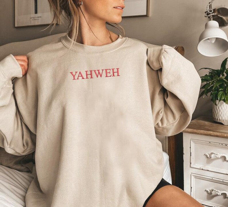 May include: A beige crewneck sweatshirt featuring the word YAHWEH embroidered in red capital letters across the center of the chest. The garment has long sleeves with ribbed cuffs and a ribbed neckline, constructed from a solid-colored fabric.