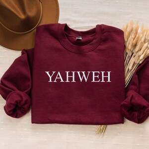 May include: A maroon long-sleeve crewneck sweatshirt featuring the word YAHWEH printed in white capital letters across the chest. The garment is displayed alongside a brown wide-brimmed hat and a bundle of dried beige ornamental grass stems.
