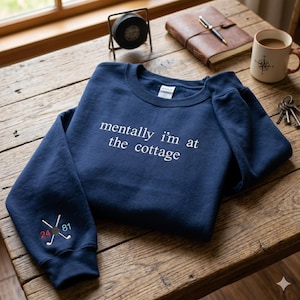 Może przedstawiać: Granatowa bluza z okrągłym dekoltem i wyhaftowanym białym napisem "mentally i'm at the cottage" na piersi. Na lewym rękawie znajduje się mała grafika przedstawiająca dwa skrzyżowane kije hokejowe, czarny krążek oraz czerwoną liczbę 24 i niebieską 81.