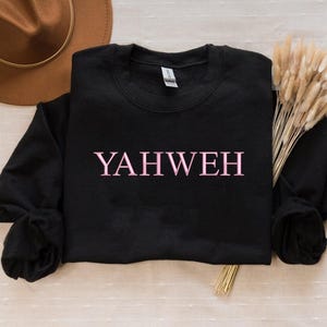 May include: A black long-sleeve crewneck sweatshirt featuring the word YAHWEH printed in light pink capital letters across the center of the chest. The garment has a ribbed neckline, cuffs, and hem, with a standard fit and a soft fabric texture.