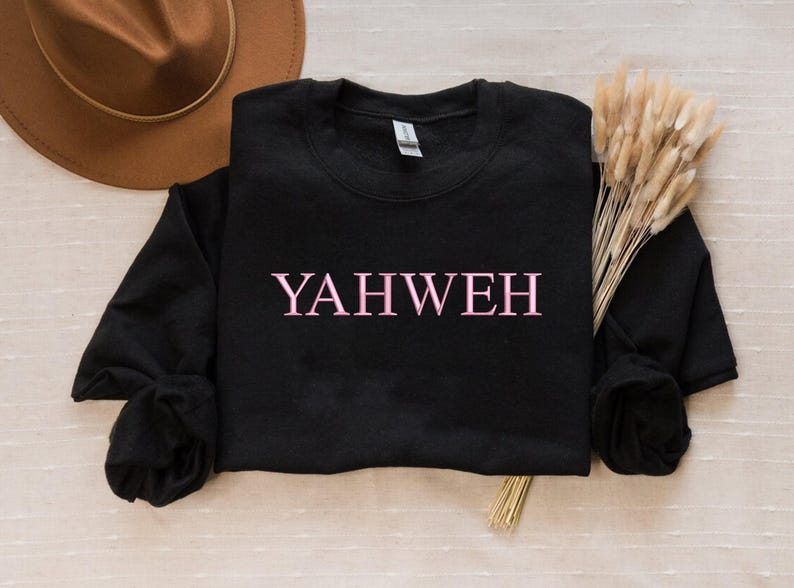May include: A black long-sleeve crewneck sweatshirt featuring the word YAHWEH printed in light pink capital letters across the center of the chest. The garment has a ribbed neckline, cuffs, and hem, with a standard fit and a soft fabric texture.