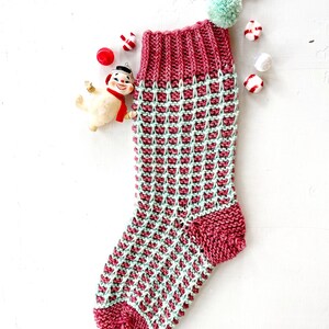 May include: A knitted Christmas stocking with a pink and green checkered pattern. The stocking has a white pom-pom on top and is decorated with a snowman ornament.