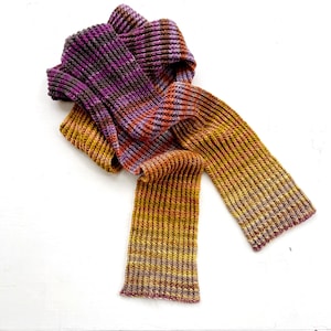 May include: A long, knitted scarf in shades of purple, orange, and brown. The scarf is folded in half and draped over a white surface.