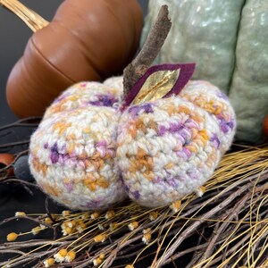 May include: A crocheted pumpkin with a white base and multi-colored yarn in shades of purple, orange, and yellow. The pumpkin is sitting on a bed of dried branches with small yellow flowers.
