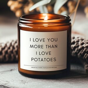 May include: A lit, amber-colored glass jar candle with a white label. The label reads "I LOVE YOU MORE THAN I LOVE POTATOES." Below the text is "natural soy candle | hand poured with love."