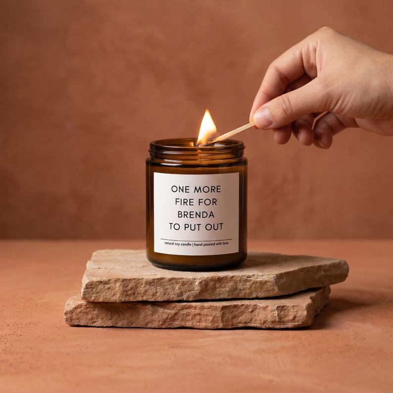 May include: A lit brown glass candle jar with a white label that reads "ONE MORE FIRE FOR BRENDA TO PUT OUT." The candle sits on two stacked, rough-hewn stone slabs. A hand lights the candle with a match.