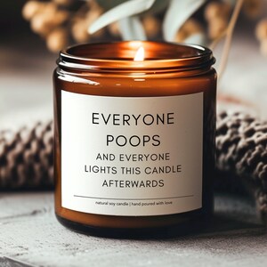 May include: A brown glass candle with a white label that reads "Everyone Poops and Everyone Lights This Candle Afterwards." The candle is lit and the flame is visible. The label also says "natural soy candle | hand poured with love."