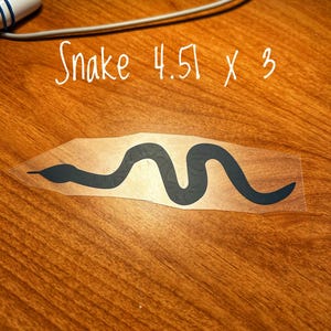 May include: A black snake decal with the text "Snake 4.51 x 3". The snake design is a simple, stylized illustration. The decal is on a clear backing, ready for application. The dimensions are 4.51 inches by 3 inches.