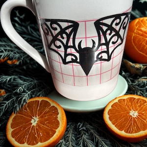 Peut inclure: Une tasse en céramique blanche ornée d'un autocollant de chauve-souris noir aux motifs d'ailes en filigrane complexes. L'autocollant est appliqué sur une feuille de transfert translucide marquée d'une grille rouge. La tasse a une anse courbée.