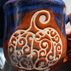 May include: A ceramic mug featuring a carved pumpkin design with intricate swirling patterns. The mug has a glossy finish with a deep blue rim that transitions into a warm, earthy brown glaze across the body and the pumpkin relief.
