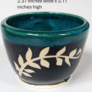 May include: A small, teal and black ceramic bowl with a white leaf design. The bowl is 2.37 inches wide and 3.11 inches high.
