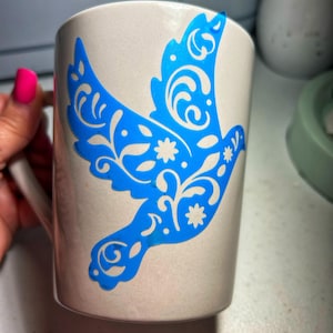May include: A light gray ceramic mug featuring a bright blue vinyl decal of a flying bird. The bird design is filled with intricate white floral and swirling patterns, creating a decorative contrast against the solid blue silhouette of the bird.