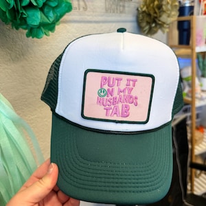 Peut inclure: Une casquette de type trucker vert foncé et blanc avec un écusson rectangulaire sur le devant. L'écusson est rose clair avec une bordure vert foncé et affiche le texte brodé "PUT IT ON MY HUSBANDS TAB" en rose, avec un petit smiley vert.