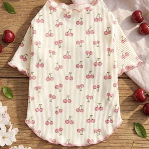 May include: Cream-colored pet shirt with a repeating pattern of pink cherries and green stems. The shirt has a ruffled neckline and sleeve edges, trimmed with pink. The ribbed fabric is soft and comfortable.