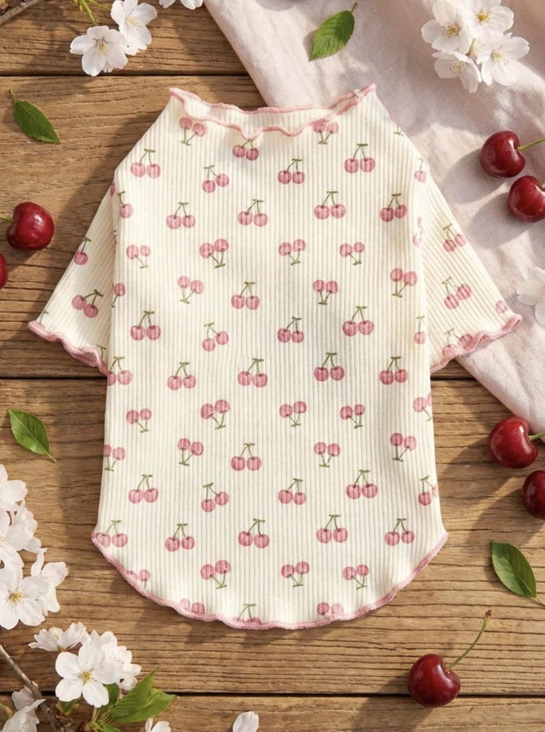 May include: Cream-colored pet shirt with a repeating pattern of pink cherries and green stems. The shirt has a ruffled neckline and sleeve edges, trimmed with pink. The ribbed fabric is soft and comfortable.
