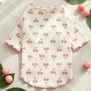 May include: Cream-colored pet shirt with a ribbed texture and a pattern of pink cherries. The shirt has short sleeves and a ruffled pink trim around the neckline and sleeves. The shirt is designed for a small dog or cat.