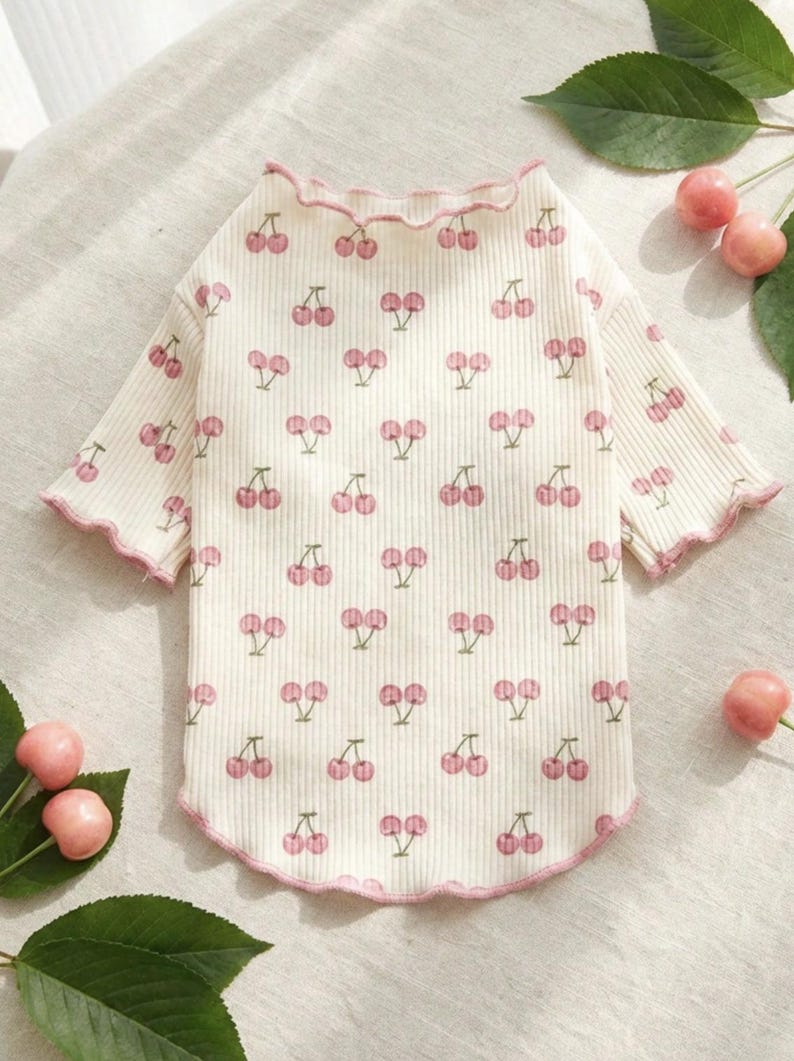 May include: Cream-colored pet shirt with a ribbed texture and a pattern of pink cherries. The shirt has short sleeves and a ruffled pink trim around the neckline and sleeves. The shirt is designed for a small dog or cat.