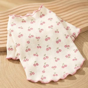 May include: White ribbed shirt with a pink cherry pattern. The shirt has a ruffled edge in pink. The cherries are pink with green stems. The shirt is laying on a wooden surface.