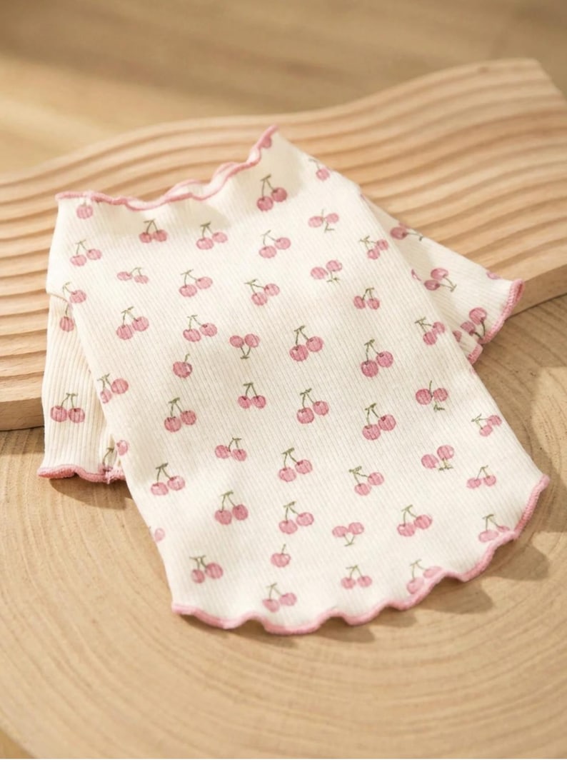 May include: White ribbed shirt with a pink cherry pattern. The shirt has a ruffled edge in pink. The cherries are pink with green stems. The shirt is laying on a wooden surface.