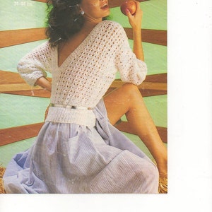 May include: A white crocheted sweater with a V-neck and a loose fit. The sweater is worn by a model with dark hair and a light blue skirt. The model is holding a red apple in her right hand. The image is from a vintage knitting pattern book.