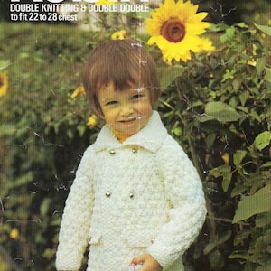 May include: A white knitted jacket with a double breasted closure and silver buttons. The jacket is being worn by a child standing in front of a green leafy background with yellow sunflowers.