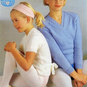 May include: Two young girls wearing white knit sweaters with a wrap design. The girl on the left is wearing a pink headband and the girl on the right is wearing a blue headband. Both girls are wearing white tights and are sitting on a white surface.