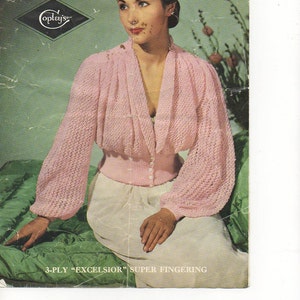 May include: A pink knitted cardigan with a V-neck and long sleeves. The cardigan has a button closure and a fitted waist. The model is wearing a white skirt and sitting on a green surface. The text "3-PLY "EXCELSIOR" SUPER FINGERING" is printed at the bottom of the image.