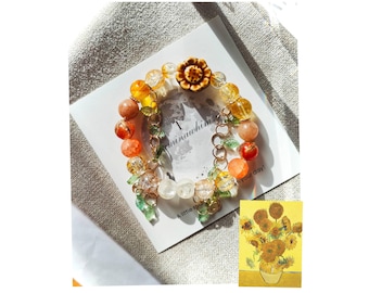 Può includere: Un braccialetto colorato con perline, un ciondolo a forma di girasole e una catenina dorata. Il braccialetto è su una carta bianca con il testo "A little something for your day!" e "T-minusawhimming".