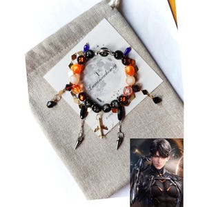 May include: A bracelet featuring orange, black, and clear beads, with silver-toned charms, including a cross and cone-shaped dangles. The bracelet is displayed on a white card with the text "Luminawhimsy" and a grey pouch.