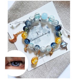 May include: A celestial-themed bracelet with blue, clear, and yellow glass beads, gold accents, and charms including a crescent moon, a star, and a globe. The bracelet is on a white card with the text "T² Luminauwhimsy" and "le sparkle to brighten your day".