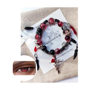 May include: A black and red beaded bracelet with a silver charm featuring a cross and a rose. The bracelet also includes a black feather charm and a silver spike charm. The bracelet is displayed on a white card with the text "T2 Luminawhimsy" and "A little to brighten your day!"