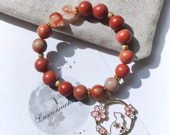 May include: A red gemstone bracelet with a gold charm featuring a white cat and pink cherry blossoms. The charm is attached to the bracelet with a gold ring. The bracelet is on a white surface with the text "T^2 Luminawhite brighten you" visible.