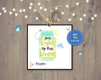 Op de afbeelding: Een wit cadeau-etiket met een blauwe en gele illustratie van een pot gevuld met vuurvliegjes. De tekst op het etiket luidt "You light up our lives!" en "From:".