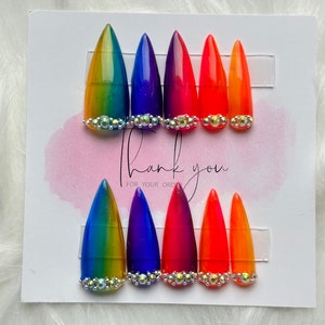 May include: Set of ten rainbow ombre press-on nails with rhinestones. The nails are in a gradient of yellow, green, blue, purple, red, and orange. The rhinestones are clear and iridescent.