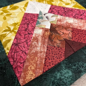 May include: A quilt block featuring a geometric design in shades of red, brown, and yellow. The block is edged with a dark green border with a floral pattern.