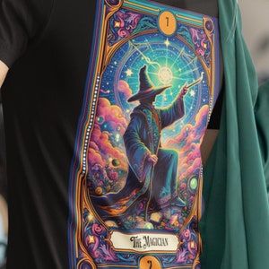 Könnte beinhalten: Ein schwarzes T-Shirt mit einer farbenfrohen Illustration einer Tarotkarte, Der Magier. Die Karte zeigt einen Zauberer in einem blauen Gewand und einem spitzen Hut, der einen Zauberstab hält und auf ein helles Licht zeigt. Die Karte ist von einem dekorativen Rahmen mit goldenen und blauen Akzenten umgeben.