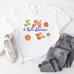 May include: White t-shirt with the text "In My Web Developer Era" in colorful retro font with flower graphics.