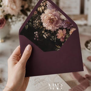 K&ouml;nnte beinhalten: Ein pflaumenfarbener Umschlag mit floralem Innendesign. Die Innenseite zeigt einen dunklen Hintergrund mit einem gro&szlig;en, detaillierten Arrangement aus rosa, lila und cremefarbenen Blumen. Der Umschlag wird ge&ouml;ffnet gehalten und zeigt das Blumenmuster.