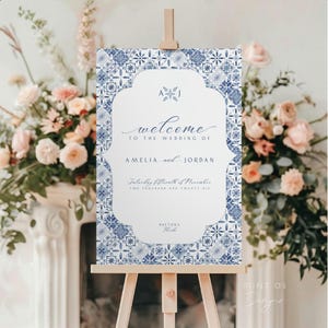 May include: A wedding welcome sign on a wooden easel. The sign features a blue and white tile pattern border with the words "welcome to the wedding of Amelia and Jordan." The date is Saturday, fifteenth of November, two thousand and twenty six, in Daytona, Florida.