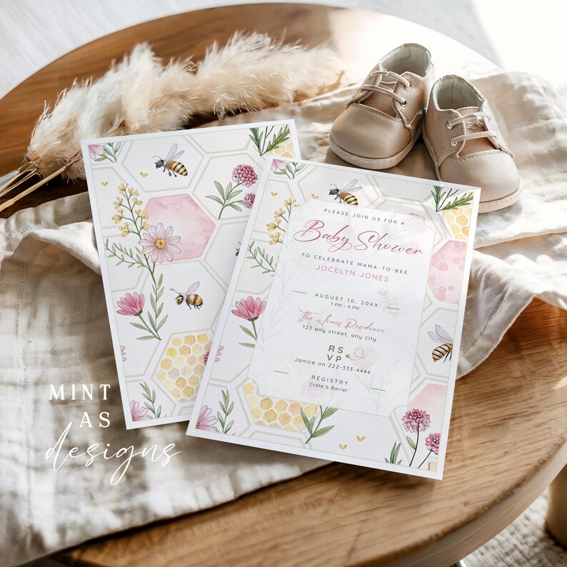 May include: Baby shower invitations with floral and bee designs. The invitations feature the text "Baby Shower" and event details. A pair of beige baby shoes rests nearby on a white cloth, with decorative pampas grass.