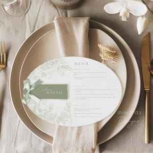 May include: Elegant table setting with a menu, place card, and gold cutlery. The menu is oval-shaped with floral designs and the word "MENU" at the top. A small green place card reads "welcome Maddie". The setting includes a fork, knife, and spoon.