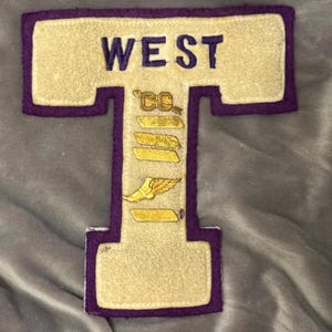 May include: A white felt letter T with a purple border. The letter is outlined in purple and has the word "WEST" above it. The letter also has a gold winged foot and the letters "CC" above it.