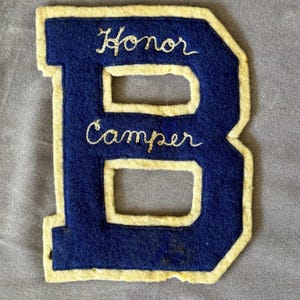 May include: Blue felt letter B with a white outline. The letter has the words "Honor Camper" embroidered in gold thread.