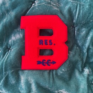 May include: Red felt letter B with blue embroidery. The letter is on a green velvet background. The embroidery says "RES." and has an arrow pointing to the right.