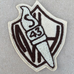 May include: A brown and beige felt patch with a shield shape. The shield has a brown outline and a white torch with the letter 'S' and the number '43' inside.