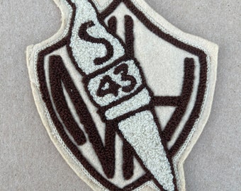 May include: A brown and beige felt patch with a shield shape. The shield has a brown outline and a white torch with the letter 'S' and the number '43' inside.