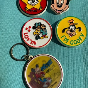May include: Five vintage Mickey Mouse themed keychains. One keychain features a red, yellow, and black star with Mickey Mouse's face in the center and the word "SUPERSTAR" written below. Another keychain features a yellow circle with Mickey Mouse's face in the center and the words "I'M GOOFY" written below. A third keychain features a white circle with Mickey Mouse and Minnie Mouse in the center and the word "LOVIN'" written below. A fourth keychain features a white circle with Mickey Mouse sitting in a chair and the words "Steamboat Willie MICKEY MOUSE" written below. The last keychain features a yellow circle with Goofy's face in the center and the words "I'M GOOFY" written below.