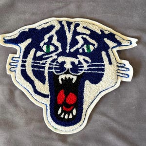 May include: A white and blue embroidered patch of a tiger's face with a red mouth and green eyes.