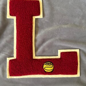 May include: A red felt letter L with a white outline and a yellow embroidered football in the lower right corner.